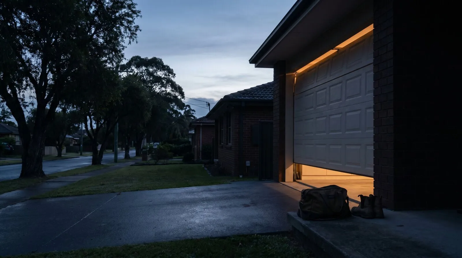 A sectional garage door mid-lift at 5:30am with warm light spilling under it onto the driveway